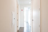 This contemporary single-floor unit offers a thoughtfully designed layout that balances open living spaces with functional private areas. The open-concept kitchen and living area serves as the heart of the unit, anchored by striking floor-to-ceiling windows that frame views of the surrounding streetscape and flood the space with natural light. The kitchen is outfitted with high-gloss white upper cabinetry, textured lower cabinets, white quartz countertops, a clean subway-tile backsplash, and a full suite of stainless steel appliances including a range, built-in microwave, dishwasher, and refrigerator. Hardwood-style flooring runs throughout the main living areas, lending warmth and continuity to the space. A well-proportioned hallway connects the living zone to the private areas of the unit, flanked by recessed panel doors and equipped with utility access. The bathroom features light tile flooring, a soaking tub with tiled surround, a wall-mounted medicine cabinet, a backlit vanity mirror, and a warm wood-toned vanity. Bedrooms are bright and airy, benefiting from large windows with street views and ample closet space. The overall aesthetic is clean, minimal, and move-in ready, with a consistent modern palette of white walls and neutral finishes throughout.