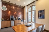 This single-floor amenity lounge is designed to serve as a versatile social hub, combining multiple recreational and functional areas within one well-appointed space. The main gathering area is anchored by a large, solid wood communal table surrounded by upholstered chairs, ideal for meetings or casual dining. Tall, arched windows trimmed in dark wood line the perimeter, flooding the room with natural light and offering views of the surrounding outdoor greenery. Crown molding and warm neutral tones give the space a refined architectural character.

Adjacent to the communal table is a dedicated billiards area, featuring a full-size pool table beneath a trio of modern, geometric pendant lights. Deep teal accent walls, a wall-mounted television, and petite bistro-style tables with contemporary seating create an inviting game room atmosphere.

A built-in coffee bar, branded as a Starbucks station, is set against a striking textured mosaic accent wall in rich, earthy tones. Warm pendant lighting and cabinetry with countertop appliances make this corner a functional refreshment hub. Completing the layout is a dedicated theater room, accessible through its own door, offering an additional entertainment option. Carpet flooring flows throughout, contributing to the overall comfort and warmth of the space.