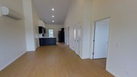 This single-floor space is defined by its striking vaulted ceilings, which rise dramatically to create an airy, expansive atmosphere throughout the open main area. The light hardwood flooring runs continuously across the space, lending warmth and visual continuity from room to room.

The kitchen is positioned toward the far end of the open area and features dark cabinetry with a contrasting navy blue island, a black refrigerator, and a sink set beneath a window that allows natural light to filter in. The dark-toned cabinetry provides a bold contrast against the bright white walls and ceiling.

Along the right-hand side of the main living area, a series of white panel doors lead to additional rooms or storage spaces, offering a well-organized and functional layout. The doors are evenly spaced and contribute to the clean, uncluttered aesthetic of the space.

The recessed lighting embedded in the vaulted ceiling ensures the interior remains well-lit throughout. The overall ambiance is open, clean, and modern, with the high ceilings and neutral palette creating a versatile environment suitable for a variety of uses.