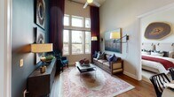 This thoughtfully designed single-floor residence makes a striking impression with its soaring ceilings and expansive windows that flood the living area with natural light. The main living space features dramatic floor-to-ceiling drapes in deep burgundy, warm hardwood flooring, and generous wall height that creates an airy, open atmosphere with views to the outdoors.

The kitchen is well-equipped with rich dark cabinetry, stainless steel appliances including a range, microwave, and refrigerator, complemented by light quartz countertops. A large center island with a secondary sink adds both function and style, while pendant lighting and elevated ceilings carry the open feel throughout.

The primary bedroom offers soaring ceilings, tall windows with patterned drapery, and direct access to a spa-inspired en suite bathroom. The primary bath features a double-sink vanity with dark cabinetry and quartz counters, a glass-enclosed tiled shower, and a spacious walk-in closet with built-in shelving. A second bedroom showcases a bold accent wall with decorative barn-door paneling and tall windows. An additional full bathroom includes a tub-shower combination, single-sink vanity with dark cabinetry, and tile flooring. Throughout the residence, the design blends contemporary and rustic elements for a warm, sophisticated ambiance.