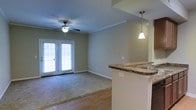 This single-floor space offers an open, flowing layout that seamlessly connects the kitchen and living areas. The kitchen features a raised bar counter topped with rich granite, providing both a functional workspace and a natural divider between the two zones. A stylish pendant light hangs above the bar, adding a warm, focused glow to the area. The living area is finished with plush carpeting, creating a comfortable, inviting atmosphere, while hardwood-style flooring defines the kitchen and entry zones. A ceiling fan with an integrated light fixture is centrally positioned in the living area, enhancing air circulation and providing ambient lighting. French doors at the far end of the living space flood the interior with natural light and offer a direct view to the outdoors, contributing to an airy, open feel. Neutral-toned walls throughout provide a versatile backdrop suited to a variety of décor styles. Crown molding adds a subtle architectural detail, lending the space a polished, finished appearance. Overall, the layout balances functionality and comfort in a well-appointed, light-filled environment.