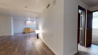 This single-floor residence offers a clean, functional layout with a cohesive design carried throughout the space. The open living area is generously proportioned, featuring warm wood-look laminate flooring, crisp white walls, and a textured ceiling. Large dark-framed windows allow natural light to filter in, creating a bright and welcoming atmosphere. The space flows seamlessly from the living area toward the kitchen, which is well-appointed with natural knotty wood cabinetry, gray laminate countertops, a peninsula with additional counter space, and white appliances including a dishwasher and refrigerator. Recessed lighting illuminates the kitchen, highlighting the ample cabinet storage above and below the counters. The bathroom is finished to match the overall aesthetic of the home, featuring the same wood-look flooring, a natural wood vanity with a gray countertop, a large wall mirror with overhead lighting, and a full soaking tub with a shower curtain rod. A built-in wall niche in the tub surround adds a practical touch. Throughout the space, consistent materials and a neutral color palette create a cohesive, move-in-ready environment suitable for a variety of uses.
