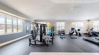 Located at 112 Mountain Flat Dr NE in Huntsville, Alabama, this fitness center offers a generously sized, single-floor layout designed to accommodate a comprehensive range of workout activities. The space features vaulted ceilings that lend an open, airy feel, complemented by recessed lighting and a ceiling fan that maintain a comfortable environment during exercise.

The main workout area is covered in durable rubber flooring and is organized into distinct training zones. A multi-station cable machine anchors the strength training section, accompanied by a flat bench and a full rack of dumbbells for free weight exercises. A dedicated accessories station displays medicine balls, resistance bands, and stability balls for functional and flexibility training.

The cardio zone is well-appointed with multiple TRUE treadmills, an Echelon smart bike with an interactive display, a recumbent bike, and a rowing machine. Wall-mounted flat-screen televisions are positioned above a large mirror, enhancing the cardio experience. Natural light streams in through several windows fitted with white blinds, creating a bright and motivating atmosphere.

An adjoining anteroom provides a transitional space connected to the main facility. The overall design is clean and modern, with neutral gray and white walls contributing to a professional and focused fitness environment.