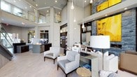 This expansive communal space makes an immediate impression with its soaring double-height ceilings and open, airy layout. The main lounge area is anchored by plush, neutral-toned sofas and accent chairs adorned with colorful throw pillows, arranged around sleek wood coffee tables that add warmth to the contemporary design. A dramatic staircase with glass railings leads to an upper mezzanine level, which overlooks the space below and adds architectural depth.

One section of the lounge features a striking stacked-stone fireplace wall, topped with a bold yellow abstract artwork that serves as a vibrant focal point. Built-in shelving flanks the fireplace, displaying curated décor pieces. Floor-to-ceiling windows flood this area with natural light and offer views of the surrounding exterior.

Toward the rear, a service counter and coffee station are visible, alongside dark wood cabinetry and display shelving, suggesting a staffed amenity area. Pendant lights in varying styles drop from the ceiling, contributing to the refined ambiance.

Large-scale glazing on one side frames views of an outdoor courtyard and pool area, seamlessly connecting the interior to the exterior. Light wood flooring runs throughout, unifying the various zones into a cohesive, upscale environment.