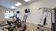 This single-floor fitness center offers a clean, functional layout designed to support a well-rounded workout routine. The space is finished with neutral-toned walls and durable, textured flooring throughout, creating a practical yet comfortable environment for exercise.

The room is generously sized, providing ample open floor space to accommodate a variety of cardio and strength training equipment. Cardio machines include multiple treadmills, elliptical trainers, a stationary bike, and a recumbent-style bike, offering users a range of options for aerobic exercise. A multi-station cable and weight machine rounds out the strength training capabilities of the space.

Several wall-mounted flat-screen televisions are positioned throughout the room, allowing users to watch content while exercising. Overhead flush-mounted lighting ensures the space is consistently well-lit. A window along one wall admits natural light, further brightening the interior.

A wood-toned entry door with a window provides access to the fitness area, while a storage unit with wicker basket drawers offers convenient organization near the entry. The fitness center connects visually to an adjacent lounge area through a full-height glass wall with dark-framed panels, offering open sightlines between the two spaces and contributing to a sense of connectivity within the broader facility.