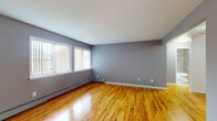 This single-floor space presents a clean, functional layout with a cohesive design aesthetic throughout. The main living areas are defined by warm-toned hardwood flooring that reflects natural light, creating a bright and inviting atmosphere. Walls are finished in a soft gray tone, complemented by crisp white trim and baseboards, lending a modern yet understated character to the space.

Large windows fitted with vertical blinds line the living area, allowing ample natural light to fill the room. Baseboard heating units are positioned along the exterior walls, providing an efficient heating solution. A ceiling fan is present in one of the rooms, adding to the functional appeal of the space.

The kitchen features light wood cabinetry paired with dark granite countertops, offering a practical contrast in materials. Stainless steel appliances, including a range, over-the-range microwave, and refrigerator, complete the kitchen setup. The floor transitions to neutral ceramic tile in this area, consistent with the practical design approach throughout.

The bathroom is well-appointed with full-wall porcelain tile surrounding the bathtub and shower combination, matching floor tile, and a wood-toned vanity topped with a dark granite countertop, accompanied by a large mirror above.
