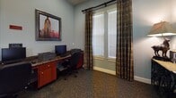 This single-floor business center is thoughtfully designed to provide a functional and comfortable workspace. The main workstation area features a continuous granite-topped desk that spans the width of the room, offering ample workspace for multiple users simultaneously. Two computer monitors and a laser printer are stationed along the desk, supported by warm-toned wood cabinetry with drawers for organized storage beneath. Ergonomic rolling office chairs complete each workstation for user comfort.

The room is finished with cool blue-gray walls that create a calm, focused atmosphere, complemented by plush, patterned carpet flooring. A large framed cityscape artwork adds a decorative focal point above the desk, infusing the space with visual interest. Natural light filters in through a sizable window dressed with elegant plaid draping curtains and white horizontal blinds, allowing for adjustable light control.

A clean, tiled corridor connects the business center to the rest of the building, featuring recessed ceiling lighting and crisp white panel doors, lending a polished and professional feel throughout. The transition between the tiled hallway and the carpeted workspace is seamless, reinforcing the space's dual focus on practicality and comfort. Overall, this business center is well-suited for guests or residents seeking a quiet, equipped environment for work or printing needs.