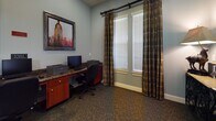 This single-floor business center is thoughtfully designed to provide a functional and comfortable workspace. The main workstation area features a continuous granite-topped desk that spans the width of the room, offering ample workspace for multiple users simultaneously. Two computer monitors and a laser printer are stationed along the desk, supported by warm-toned wood cabinetry with drawers for organized storage beneath. Ergonomic rolling office chairs complete each workstation for user comfort.

The room is finished with cool blue-gray walls that create a calm, focused atmosphere, complemented by plush, patterned carpet flooring. A large framed cityscape artwork adds a decorative focal point above the desk, infusing the space with visual interest. Natural light filters in through a sizable window dressed with elegant plaid draping curtains and white horizontal blinds, allowing for adjustable light control.

A clean, tiled corridor connects the business center to the rest of the building, featuring recessed ceiling lighting and crisp white panel doors, lending a polished and professional feel throughout. The transition between the tiled hallway and the carpeted workspace is seamless, reinforcing the space's dual focus on practicality and comfort. Overall, this business center is well-suited for guests or residents seeking a quiet, equipped environment for work or printing needs.