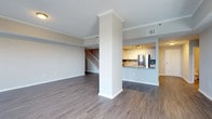 This two-storey space offers a thoughtfully designed open-concept main level that balances functionality and modern aesthetics. The living area is expansive and airy, featuring smooth hardwood-style flooring in a warm grey-toned finish that runs throughout the main level, complemented by crisp white crown moulding and neutral grey walls that create a clean, contemporary atmosphere.

A structural column with decorative moulding serves as a subtle architectural divider between the living area and the kitchen pass-through. The kitchen is partially open to the main living space via a raised breakfast bar with a stone or granite countertop, providing both a visual connection and a casual dining option. White upper cabinetry, a stainless steel refrigerator, track lighting, and a tile backsplash complete the kitchen's modern look.

A staircase with white balusters and a wooden railing leads to the upper floor, contributing to the vertical openness of the space. Recessed ceiling vents and neutral finishes throughout ensure the space feels well-ventilated and move-in ready. Overall, the main level exudes a bright, functional, and contemporary character.