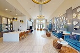 Located at 650 Sgt Ed Holcomb Blvd in Conroe, TX, this expansive single-floor community clubhouse offers a generously proportioned, thoughtfully designed interior that blends modern elegance with comfortable functionality. The grand main lounge commands attention with its dramatic vaulted ceilings adorned with tiered gold chandelier rings and recessed lighting, all set above large-format light stone tile flooring. A striking feature wall of varied geometric gold-framed mirrors anchors a plush seating area furnished with rich blue velvet sofas, tufted caramel leather ottomans, and gold-accented chairs, creating a sophisticated yet inviting atmosphere. A long marble-topped counter with warm leather seating serves as a stylish workspace or leasing area along one side of the room. Floor-to-ceiling glass entry doors flood the space with natural light and offer views to the outdoors. A separate lounge zone features a dramatic tall upholstered banquette booth beneath an illuminated architectural panel, surrounded by additional seating clusters. A private business center or flex room provides a dedicated work area with a desk, upholstered chairs, and large windows overlooking the exterior grounds. A private conference or meeting room with a round wood table, leather chairs, and a bold octagonal gold pendant light completes the offering, making this clubhouse a well-appointed hub for community life.