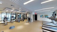 This fitness center offers a well-organized and spacious workout environment designed to accommodate a range of exercise needs. The space features a drop ceiling with recessed linear fluorescent lighting that provides consistent, even illumination throughout. The flooring consists of durable, cushioned rubber-like material in a neutral tan tone, suitable for both cardio and strength training activities.

The equipment layout includes a cable cross machine, a multi-station weight trainer, an elliptical trainer, and a stair climber, all positioned to allow comfortable circulation between stations. Along the far right side of the space, a row of treadmills lines the wall, maximizing floor usage efficiently. A modular shelving unit serves as a divider between workout zones, adding subtle organization to the layout.

The walls are white and largely unadorned, contributing to a clean, clinical aesthetic. A single piece of colorful wall art adds a subtle decorative touch near the exit door. Large windows near the entrance allow natural light to filter in, brightening the front of the space. The overall ambiance is functional, modern, and well-maintained, making it a practical facility for daily fitness use.