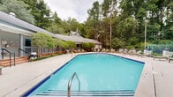 Located at 10 Arbor Crossing Dr in Lithonia, Georgia, this outdoor amenity area serves as a welcoming recreational retreat set against a backdrop of mature trees and natural Georgia greenery. At the heart of the space is a generously sized swimming pool with clear turquoise water, framed by a wide concrete sundeck lined with cushioned lounge chairs and chaise lounges, offering ample space for relaxation and sunbathing. A separate hot tub or wading pool area is situated adjacent to the main pool, marked with safety signage and accessible via a dedicated entry stairway. The pool area is enclosed by a secure metal fence with decorative column posts, ensuring safety while maintaining an open, airy feel. A well-maintained clubhouse or pool house building anchors one side of the deck, featuring a gabled roof with a dormer window, a covered porch with railings, and manicured shrubs along its facade. On the opposite side of the pool, a playground structure is visible, adding family-friendly functionality to the space. Seating areas with outdoor furniture, including sofas and accent chairs, are thoughtfully arranged around the perimeter, creating comfortable gathering spots. The entire area is surrounded by tall trees that provide natural shade and privacy, enhancing the overall sense of a serene, resort-like outdoor environment.
