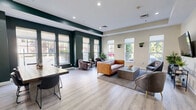 Located at 73 Brownstone Way in Englewood, NJ, this well-appointed communal lounge offers a thoughtfully designed single-floor space that balances comfort, style, and functionality. The open-plan layout is anchored by light-toned hardwood flooring and a tray ceiling with recessed lighting, creating a bright and welcoming atmosphere throughout.

A bold deep-green accent wall frames one side of the room, providing a striking contrast against the warm beige tones of the remaining walls. Multiple large windows line the perimeter, flooding the space with generous natural light and offering views of the lush greenery outside. Wall-mounted planters add a fresh, organic touch to the décor.

The lounge is divided into distinct functional zones. A comfortable seating area features a cognac leather sofa, leather accent chairs, and a concrete-topped coffee table arranged atop a neutral area rug — ideal for relaxed conversation or casual gatherings. A large wall-mounted flat-screen television with a media console anchors a dedicated viewing area nearby.

A dining and work zone accommodates a solid wood communal table with seating, positioned beside a white credenza with countertop amenities. Framed artwork decorates the walls, adding a curated, residential feel. French doors provide access to adjacent facilities, completing this versatile and inviting community space.