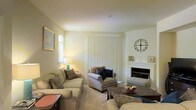 This single-floor residence offers a welcoming living area defined by its warm, neutral color palette and thoughtfully arranged furnishings. The living room serves as the heart of the space, centered around a classic white-mantled fireplace that provides a natural focal point and adds a sense of warmth and character to the room. Plush, neutral-toned sofas and accent chairs are arranged to encourage conversation and comfort, complemented by a dark wood coffee table that anchors the seating area.

Natural light filters into the room through an arched side window, while a set of vertical blinds covering a large sliding or patio door allows for adjustable brightness throughout the day. The soft, carpeted flooring enhances the cozy ambiance, and subtle decorative touches — including framed artwork, a decorative wall clock, and tasteful accessories on the mantle — contribute to an inviting, curated atmosphere. The cream-toned walls reflect light effectively, keeping the space feeling open and airy. Overall, the living area strikes a balance between functional comfort and understated elegance.