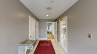 The Macomb Manor fitness and amenity center is a single-floor facility offering a practical and well-organized layout across a comfortable footprint. The space is entered through a welcoming corridor finished with light tile flooring and branded Macomb Manor entry rugs, setting a clean, professional tone. This hallway connects key areas of the facility and includes a water fountain, restroom access, and a utility sink, ensuring convenience for all users.

The fitness area is divided into two connected zones, both carpeted for comfort during exercise. The first zone features cardio equipment, including stationary bikes and other machines, complemented by a full-length wall mirror that enhances the sense of space. French doors with glass panes line one wall, allowing natural light to filter in and providing views of the exterior. The second fitness zone houses a multi-station strength training machine alongside additional cardio equipment, with fluorescent overhead lighting ensuring a well-lit workout environment. The gray-toned walls throughout maintain a neutral, focused atmosphere. Together, these spaces form a functional and accessible amenity center suited to everyday fitness needs.