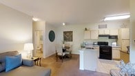 This single-floor residence is designed around an open-concept floor plan that integrates the kitchen, dining, and living areas into one cohesive, flowing space. The kitchen is outfitted with light wood cabinetry, a black electric range, and a built-in sink set within a white peninsula counter that doubles as a natural divider between the kitchen and the rest of the living space. Tile flooring anchors the kitchen zone, while plush carpeting extends through the dining and living areas, adding warmth underfoot. The dining area comfortably accommodates a round table with upholstered chairs and is accented by decorative wall art, contributing to the space's understated charm. A ceiling fan overhead provides both light and airflow to the central living zone. The living area connects seamlessly, offering ample room for seating and relaxation. A hallway visible from the main area leads to a bathroom, hinting at a thoughtfully laid-out floor plan. Neutral wall tones and consistent finishes throughout create a calm, cohesive ambiance well-suited for everyday living.