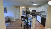 This single-floor residence offers a well-designed, open-concept layout that seamlessly connects the kitchen and living areas. The kitchen is a standout feature, showcasing crisp white cabinetry that extends to the upper walls, providing generous storage space. Dark countertops offer a striking contrast against the white cabinets, while a central island with a matching dark countertop doubles as a breakfast bar, complete with seating for two. Pendant lighting above the island adds a warm, focused glow to the workspace. Black appliances, including a refrigerator, microwave, and range, complement the kitchen's bold color palette. A decorative diamond-pattern wine rack adds a charming architectural detail above the pass-through area. Rich, dark hardwood-style flooring runs throughout the space, grounding the open layout with warmth and continuity. A textured ceiling adds subtle character overhead. The open sightlines from the kitchen into the adjacent rooms create an inviting, connected flow, making the space feel airy and functional. Natural light filters in from neighboring rooms, enhancing the overall brightness and welcoming ambiance of this thoughtfully designed home.
