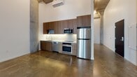 This single-floor loft showcases a compelling blend of industrial character and modern refinement. The open-plan layout is anchored by polished concrete floors that span the entire space, reflecting light and reinforcing the unit's clean, contemporary aesthetic. Soaring ceilings with exposed concrete soffits and structural columns lend a raw, warehouse-inspired atmosphere, while crisp white walls provide a bright and neutral contrast.

The kitchen is thoughtfully designed, featuring warm walnut-toned cabinetry extending floor to ceiling, a light-toned subway tile backsplash, and sleek light countertops. Stainless steel appliances — including a refrigerator, wall oven, microwave, dishwasher, and an induction cooktop — deliver a fully equipped culinary setup. Under-cabinet lighting adds a polished finishing touch.

The living area is generously proportioned, with a prominent concrete column as a striking architectural focal point. A glass sliding door with sheer curtains fills the space with natural light and provides direct access to an outdoor area.

The bathroom features classic white subway tile surrounding the soaking tub and shower combination, with brushed nickel grab bars, a toilet, and a floating vanity — all set on matching concrete flooring that ties cohesively to the rest of the unit. A wall-mounted mini-split HVAC system ensures year-round comfort throughout this distinctive space.