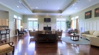 Vintage is a thoughtfully designed single-floor community space that exudes warmth and classic elegance. The centerpiece of the space is a spacious lounge area anchored by a traditional fireplace, which serves as a natural gathering point and adds a welcoming ambiance to the room. Rich, polished hardwood floors stretch throughout, reflecting the natural light that floods in through large French doors and transom windows lining the walls.

The lounge is generously appointed with comfortable seating arrangements, including sofas, armchairs, and accent tables, creating multiple distinct conversation areas suited for both socializing and quiet relaxation. A wall-mounted flat-screen television above the fireplace adds a modern touch to the otherwise traditionally styled interior.

The soft sage-green walls, accented by crisp white crown molding and wainscoting, lend the space a refined yet approachable character. Tray ceilings with recessed lighting enhance the sense of height and openness, while potted greenery and warm drapery introduce organic texture and color throughout.

The French doors offer views of lush outdoor greenery, seamlessly connecting the interior to the natural surroundings. Overall, Vintage presents a harmonious blend of classic architectural details and comfortable furnishings, making it an inviting and functional communal environment.