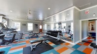 This fitness center is designed to support a comprehensive workout experience, combining functionality with an energetic aesthetic. The space is outfitted with a variety of cardio equipment, including treadmills, an elliptical, and a rowing machine, providing members with multiple options for aerobic training. A full dumbbell rack offers a wide range of free weights for strength training, complemented by adjustable weight benches positioned throughout the floor.

The interior features light gray walls accented by crisp white crown molding, creating a clean and polished backdrop. A large wall-to-wall mirror reflects the space, enhancing the sense of openness while allowing users to monitor their form during workouts. Recessed lighting and ceiling fans ensure the area remains well-lit and comfortably ventilated throughout use.

The flooring makes a bold visual statement with a vibrant, multi-colored tile pattern featuring shades of orange, teal, blue, and gray — adding a dynamic, motivating energy to the environment. Natural light filters in through multiple windows, further brightening the space. Safety signage and informational notices are posted near the entrance, and a wood-paneled entry door adds a warm, welcoming touch to the overall design.