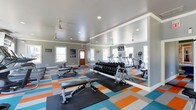 This fitness center is designed to support a comprehensive workout experience, combining functionality with an energetic aesthetic. The space is outfitted with a variety of cardio equipment, including treadmills, an elliptical, and a rowing machine, providing members with multiple options for aerobic training. A full dumbbell rack offers a wide range of free weights for strength training, complemented by adjustable weight benches positioned throughout the floor.

The interior features light gray walls accented by crisp white crown molding, creating a clean and polished backdrop. A large wall-to-wall mirror reflects the space, enhancing the sense of openness while allowing users to monitor their form during workouts. Recessed lighting and ceiling fans ensure the area remains well-lit and comfortably ventilated throughout use.

The flooring makes a bold visual statement with a vibrant, multi-colored tile pattern featuring shades of orange, teal, blue, and gray — adding a dynamic, motivating energy to the environment. Natural light filters in through multiple windows, further brightening the space. Safety signage and informational notices are posted near the entrance, and a wood-paneled entry door adds a warm, welcoming touch to the overall design.