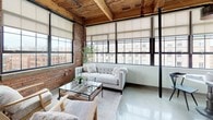 This 1-bedroom, 1-bathroom urban loft masterfully blends industrial character with modern comfort. The living area is a standout, featuring dramatic exposed brick walls, soaring reclaimed wood-plank ceilings with heavy timber beams, and a bank of large black-framed industrial windows that flood the space with natural light and frame expansive city views. Polished concrete floors run throughout, tying the open layout together seamlessly.

The kitchen is thoughtfully designed with grey shaker cabinetry, granite-style countertops, and stainless steel appliances, including a French-door refrigerator and built-in microwave. A peninsula with an extended breakfast bar and metal stools provides casual seating and defines the kitchen from the living space. Pendant lighting, including white dome and ornate metallic fixtures, adds character overhead.

The bedroom offers a cozy retreat with exposed beams overhead, a large window for natural light, and a wall-mounted mini-split system for year-round comfort. A small entry nook with exposed brick and wood-framed glass doors adds a charming, intimate corner to the home. The bathroom is clean and functional, featuring a tub-shower combo, a granite-topped vanity with white cabinetry, and a wood-framed mirror.