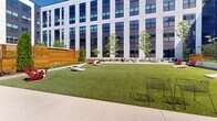 Located at 877 Lavina St in Fort Wayne, Indiana, this outdoor courtyard is a thoughtfully designed communal space nestled within the interior of a contemporary multi-story building. The building's modern façade — featuring a bold combination of dark panel cladding and white structural framing alongside large windows — encloses the courtyard on multiple sides, creating a sheltered yet open-air environment filled with natural light.

At the heart of the courtyard lies a well-maintained turf lawn, ideal for recreational activities such as lawn games, including cornhole boards visible on-site. The lawn is bordered by wide concrete walkways that connect the various zones of the space and provide direct access to the building's interior through multiple entry points.

Landscaped garden beds line the perimeter, featuring young deciduous trees, flowering shrubs, and mulched plantings that introduce natural greenery and soften the architectural surroundings. A dedicated patio area offers an assortment of seating configurations, including lounge chairs, accent tables, and umbrella-shaded spots — ideal for casual gatherings or individual relaxation. String lights are strung overhead in portions of the courtyard, adding a welcoming ambiance. A warm-toned horizontal wood screen wall creates a defined, semi-private nook within the space. The overall design balances functionality with aesthetic appeal, making it a versatile outdoor amenity.