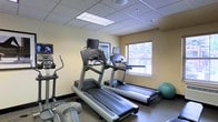 This fitness room is a dedicated exercise space designed to support cardio-focused workouts. The room features multiple treadmills equipped with individual monitor screens, allowing users to enjoy entertainment while exercising. An elliptical machine is positioned along one wall, complementing the treadmill options and providing low-impact cardio alternatives. A stability ball is also present, adding versatility for core and stretching exercises.

The space is finished with a drop tile ceiling fitted with recessed fluorescent lighting panels, ensuring bright, even illumination throughout. The walls are painted in a neutral warm tone, contributing to a clean and functional atmosphere. A window fitted with horizontal blinds allows natural light to filter into the room, enhancing the overall environment.

The flooring is a durable, dark-toned surface suited for high-traffic exercise use. The layout is efficient, with equipment arranged to maximize usability within the available footprint. A framed piece of wall art adds a modest decorative touch. Overall, the fitness room is practical and well-equipped for essential cardiovascular training needs.