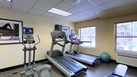 This fitness room is a dedicated exercise space designed to support cardio-focused workouts. The room features multiple treadmills equipped with individual monitor screens, allowing users to enjoy entertainment while exercising. An elliptical machine is positioned along one wall, complementing the treadmill options and providing low-impact cardio alternatives. A stability ball is also present, adding versatility for core and stretching exercises.

The space is finished with a drop tile ceiling fitted with recessed fluorescent lighting panels, ensuring bright, even illumination throughout. The walls are painted in a neutral warm tone, contributing to a clean and functional atmosphere. A window fitted with horizontal blinds allows natural light to filter into the room, enhancing the overall environment.

The flooring is a durable, dark-toned surface suited for high-traffic exercise use. The layout is efficient, with equipment arranged to maximize usability within the available footprint. A framed piece of wall art adds a modest decorative touch. Overall, the fitness room is practical and well-equipped for essential cardiovascular training needs.