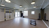 This fitness studio offers a clean, functional space designed to support a wide range of workout activities. The open floor plan is finished with smooth, light-colored flooring that provides a safe and comfortable surface for exercise, stretching, and movement-based training. The room is generously illuminated by a combination of recessed circular ceiling fixtures and natural light streaming in through large windows and glass-paneled double doors, creating a bright and energizing atmosphere. Exposed piping along the ceiling adds an industrial character to the otherwise neutral, light-gray interior. A TRX suspension training system is mounted near the entrance, ideal for bodyweight strength and conditioning exercises. The space also includes a stability ball and a plyo box, offering versatile options for cardio, strength, and functional fitness routines. Wall-mounted instructional charts provide guidance for workouts. The double doors open to an exterior area, allowing for excellent ventilation and easy access. Overall, the studio presents a practical and inviting environment well-suited for personal training sessions, group fitness classes, or individual workouts.