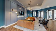 This well-appointed clubroom offers a sophisticated and inviting atmosphere designed for both socializing and relaxation. The space is defined by a bold teal-blue color palette that spans the walls throughout, complemented by rich hardwood-style flooring and crisp white crown molding that adds a polished finish.

The central lounge area serves as the heart of the room, featuring a pair of cognac leather sofas arranged around an industrial-style coffee table, all anchored by a textured area rug. A panel-accented feature wall in muted slate gray frames a wall-mounted flat-screen TV, flanked by articulating brass and matte black sconce lighting. Overhead, a statement geometric chandelier adds a modern, upscale touch.

Adjacent to the lounge, a full kitchen is outfitted with dark navy cabinetry, a subway tile backsplash, stainless steel appliances, and a generous island topped with a marble-veined countertop and lined with black bar stools — ideal for casual dining or entertaining. Pendant lights hang above the island, enhancing both function and style.

A dedicated dining nook accommodates a solid wood table with modern cantilever chairs, bathed in natural light from a trio of large windows dressed with wood blinds and charcoal curtain panels. French doors provide access to the exterior, further brightening the space and offering a seamless indoor-outdoor connection.