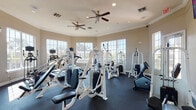 This fitness center is a thoughtfully designed, single-floor facility built to support a full range of workout routines. The space is bright and open, benefiting from an abundance of large, multi-pane windows that line multiple walls, flooding the interior with natural light and offering pleasant views of surrounding palm trees and greenery. The warm, neutral-toned walls are accented by crown molding, lending the room a polished, well-maintained aesthetic. Underfoot, dark rubber flooring provides both durability and safety throughout the workout area.

The strength training section is outfitted with a comprehensive array of Hoist-brand cable and selectorized machines, covering a wide variety of muscle groups. These are complemented by additional weight machines suited for both upper and lower body training. The cardio section features multiple SportsArt treadmills, each equipped with individual display monitors, as well as a stationary upright cycle. A wall-mounted flat-screen television is positioned for easy viewing during workouts. Overhead, dual ceiling fans ensure consistent air circulation, enhancing comfort during exercise. The facility is also clearly marked with exit signage and is accessible via a glass-paneled entry door, reflecting attention to both functionality and safety standards.