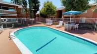 This outdoor recreational area centers around a generously sized swimming pool with clear, blue-green water, complemented by a separate circular spa adjacent to it. The pool deck is finished in a warm terracotta-toned surface, providing ample space for lounging and socializing. Multiple cushioned lounge chairs are arranged around the pool, alongside several seating areas furnished with chairs and tables, each shaded by vibrant blue market umbrellas that offer relief from the sun. The entire area is enclosed by a white metal security fence with a gated entry, ensuring a safe and private environment. A poolside utility area features French doors with louvered shutters and an outdoor shower, offering convenient access and rinse-off facilities for swimmers. Mature trees line the perimeter, providing natural shade and a lush green backdrop that softens the surrounding building facades. The multi-story residential buildings, constructed with earthy brown stucco and accented with wood detailing and balconies, frame the courtyard on multiple sides. The space is well-maintained, bright, and designed to maximize outdoor enjoyment under open skies, making it an inviting retreat for relaxation and recreation.