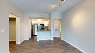 This single-floor unit offers a thoughtfully designed open-concept layout that maximizes both functionality and visual appeal. The living area features warm hardwood-style flooring that extends throughout the main living space, creating a cohesive and inviting atmosphere. A textured ceiling adds dimension overhead, while neutral gray-toned walls provide a clean, contemporary backdrop throughout.

The kitchen is a standout feature, anchored by a peninsula with granite-look countertops that serves as both a prep area and casual dining space. White cabinetry provides ample storage, complemented by a tasteful tile backsplash and a full suite of stainless steel appliances, including a refrigerator, microwave, and range. Recessed track lighting highlights the kitchen area, adding both style and practicality.

The open flow between the kitchen and living area makes the space feel expansive and well-connected. A hallway leads to additional rooms, including what appears to be a bathroom, offering convenient accessibility. Multiple doors suggest well-defined private spaces beyond the main living area. HVAC vents are strategically placed for efficient climate control. Overall, the unit balances modern finishes with a functional layout, making it a comfortable and well-appointed living space.