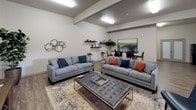 This single-story clubhouse is designed to serve as a welcoming community hub, offering a thoughtfully arranged open-concept layout with distinct functional zones that flow seamlessly into one another. The interior features durable wood-style flooring throughout, neutral-toned walls, and recessed ceiling details that add subtle architectural interest while maintaining a clean, contemporary aesthetic.

The main lounge area is generously proportioned, accommodating multiple seating arrangements including large gray sofas, accent chairs, and a tufted ottoman coffee table centered on a patterned area rug. Decorative wall art and floating shelves add visual character without cluttering the space. Adjacent to the lounge is a dedicated media area equipped with a large flat-screen television mounted on a barn-door-style entertainment console, complemented by additional seating for casual viewing.

A dining nook with a high-top table and bar-style seating provides a functional space for small group gatherings or work sessions. Toward the entry, glass-paneled double doors flood the foyer with natural light and offer a direct view to the outdoor courtyard beyond. The overall ambiance is modern, comfortable, and community-oriented, balancing open social space with quieter, more intimate corners.