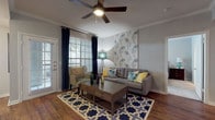 This single-story, two-bedroom, two-bathroom residence features a cohesive design characterized by warm wood-style flooring, crown molding, and neutral gray walls throughout the main living areas. The living room is a comfortable, well-lit space enhanced by a ceiling fan, large windows with access to an outdoor area, and striking accent wall detailing that adds visual interest.

Adjacent to the living room, the dining area benefits from natural light streaming through a sizable window and features a tray ceiling with a pendant light fixture overhead. A decorative botanical wall panel serves as a focal point in this space, which opens conveniently toward the kitchen.

The kitchen is well-equipped with white cabinetry, stainless steel appliances including a refrigerator, range, and microwave, complemented by a gray subway tile backsplash and light countertops. Wood-style flooring continues throughout, reinforcing a cohesive aesthetic.

Both bedrooms are generously sized, carpeted for comfort, and receive ample natural light through large windows. The primary bedroom connects to an en-suite bathroom featuring a glass-enclosed shower, wood-style flooring, and access to a walk-in closet. The second bathroom offers a full tub-shower combination with gray cabinetry and white countertops.