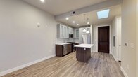 This well-designed single-floor space showcases a clean, contemporary aesthetic throughout. The open-concept kitchen serves as the heart of the layout, combining functionality with modern style. It features white upper cabinetry with frosted glass-front doors, contrasted by rich dark-wood lower cabinets that add warmth and depth. A full suite of stainless steel appliances — including a refrigerator, dishwasher, and range — lines the walls, complemented by a classic white subway tile backsplash. A central kitchen island with a crisp white countertop provides additional prep space and a natural gathering point, illuminated by two elegant pendant lights hanging overhead. A skylight positioned above the kitchen floods the space with natural daylight, creating a bright and airy atmosphere. Light wood-toned flooring runs throughout, tying the space together with a warm, neutral base. Recessed lighting adds to the well-lit environment, while the neutral gray walls provide a versatile backdrop. A dark-stained arched wood door adds a touch of character and architectural contrast to the otherwise modern design, giving the space a unique and inviting feel.