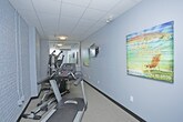This single-floor fitness space is designed to provide a functional and efficient workout environment. The room features painted white brick walls that lend a clean, modern aesthetic, complemented by a drop ceiling with flush-mounted lighting that ensures the space is well-lit throughout. A large wall-mounted mirror expands the sense of space and allows users to monitor their form during exercise. The room is equipped with cardio machines, including a treadmill prominently positioned in the foreground, alongside additional strength and multi-use training equipment visible in the mirrored reflection. A wall-mounted mini-split air conditioning unit provides dedicated climate control, keeping the environment comfortable during workouts regardless of the season. Decorative artwork on the walls adds a subtle touch of personality to the otherwise utilitarian space. The flooring is designed to support gym use, providing a practical surface suitable for a variety of exercises. Overall, the space offers a focused and well-maintained fitness facility with all the essential elements needed for a complete workout routine.
