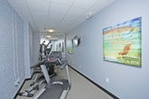 This single-floor fitness space is designed to provide a functional and efficient workout environment. The room features painted white brick walls that lend a clean, modern aesthetic, complemented by a drop ceiling with flush-mounted lighting that ensures the space is well-lit throughout. A large wall-mounted mirror expands the sense of space and allows users to monitor their form during exercise. The room is equipped with cardio machines, including a treadmill prominently positioned in the foreground, alongside additional strength and multi-use training equipment visible in the mirrored reflection. A wall-mounted mini-split air conditioning unit provides dedicated climate control, keeping the environment comfortable during workouts regardless of the season. Decorative artwork on the walls adds a subtle touch of personality to the otherwise utilitarian space. The flooring is designed to support gym use, providing a practical surface suitable for a variety of exercises. Overall, the space offers a focused and well-maintained fitness facility with all the essential elements needed for a complete workout routine.