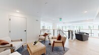 This generously sized single-floor amenity space is thoughtfully designed to offer both social and leisure experiences. The layout encompasses a hallway and a expansive living area that flows across a wide, open footprint.

The living lounge area is bright and airy, finished with wide-plank light hardwood flooring and crisp white walls that maximize the sense of space. Large windows line the perimeter, flooding the room with abundant natural light and providing open views of the surrounding greenery. A cable-railed mezzanine overlooks the floor below, adding architectural depth and a sense of vertical openness. The lounge features multiple seating arrangements — including mid-century modern armchairs in neutral tones and plush sofas — anchored by area rugs and a statement chandelier. An elevator provides convenient access, and curated wall art adds a welcoming, residential touch.

The screening room offers a striking contrast in atmosphere, enveloped in deep blue-gray tones with a coffered ceiling and arched wall niches fitted with elegant sconce lighting. Rich cognac leather seating — including a long sectional and tufted lounge chairs — is arranged theater-style facing a wall-mounted display screen, creating an intimate and upscale cinematic experience. Together, these spaces deliver a polished, amenity-rich environment designed for both relaxation and entertainment.