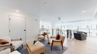 This generously sized single-floor amenity space is thoughtfully designed to offer both social and leisure experiences. The layout encompasses a hallway and a expansive living area that flows across a wide, open footprint.

The living lounge area is bright and airy, finished with wide-plank light hardwood flooring and crisp white walls that maximize the sense of space. Large windows line the perimeter, flooding the room with abundant natural light and providing open views of the surrounding greenery. A cable-railed mezzanine overlooks the floor below, adding architectural depth and a sense of vertical openness. The lounge features multiple seating arrangements — including mid-century modern armchairs in neutral tones and plush sofas — anchored by area rugs and a statement chandelier. An elevator provides convenient access, and curated wall art adds a welcoming, residential touch.

The screening room offers a striking contrast in atmosphere, enveloped in deep blue-gray tones with a coffered ceiling and arched wall niches fitted with elegant sconce lighting. Rich cognac leather seating — including a long sectional and tufted lounge chairs — is arranged theater-style facing a wall-mounted display screen, creating an intimate and upscale cinematic experience. Together, these spaces deliver a polished, amenity-rich environment designed for both relaxation and entertainment.