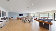 This contemporary single-floor community lounge is designed for both relaxation and social engagement, offering a generously open layout bathed in natural light. Large floor-to-ceiling windows with black-framed sliding glass doors line the exterior wall, providing sweeping views of lush greenery and direct access to an outdoor terrace furnished with bar-height tables and seating.

The interior is anchored by a comfortable lounge area featuring a warm-toned sectional sofa, modern accent chairs in cognac leather and gray upholstery, and a centrally placed coffee table — all arranged to encourage conversation and relaxation. A wall-mounted flat-screen TV is set against a soft blue-gray feature wall, adding a subtle pop of color to the otherwise crisp white palette.

Entertainment is well catered for with a full-size pool table and additional game table positioned near the windows, alongside bar stool seating for casual gathering. A long conference-style dining table with vibrant blue chairs provides a dedicated space for group meetings or communal dining.

A built-in utility area with sleek, high-gloss gray cabinetry and an integrated sink serves practical needs. Light wood flooring flows throughout, unifying the space and enhancing its bright, welcoming ambiance. A decorative shelving wall and double French doors complete the sophisticated, functional design.