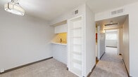 This single-floor residential space is thoughtfully designed with a clean, neutral aesthetic throughout. The layout includes multiple carpeted bedrooms, each finished with soft neutral-toned carpet, ceiling fans with integrated lighting, and windows fitted with blinds that allow natural light to filter in while maintaining privacy. The rooms are generously proportioned, offering ample space for flexible furniture arrangements. At least one bedroom features a closet with a hanging rod for convenient storage.

A standout feature of the space is a built-in wet bar area, accented by white painted brick detailing along the base, a countertop with a modern gold-toned faucet, and an adjacent floor-to-ceiling open bookshelf unit. This area adds both functionality and visual interest, making it a versatile nook for entertaining or additional storage.

The bathroom is well-appointed with wood-look tile flooring, a full soaking tub and shower combination with white subway tile surround, a white vanity with quartz-style countertop, brushed nickel fixtures, and a warm wood-framed mirror above. The overall color palette across the space is light and airy, with white walls, dark baseboards, and consistent finishes that create a cohesive and inviting atmosphere throughout.