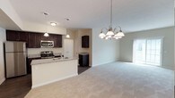 This single-floor residence offers a thoughtfully designed open-concept layout that seamlessly connects the kitchen, dining, and living areas. The kitchen is a standout feature, showcasing rich dark cabinetry, granite countertops, and a full suite of stainless steel appliances including a refrigerator, electric range, and built-in microwave. A peninsula with a sink provides additional counter space and serves as a natural divider between the kitchen and the expansive carpeted living area, which is illuminated by an elegant multi-arm chandelier. Sliding glass doors in the living area invite natural light and provide access to the outdoors. The primary bedroom is generously sized with plush carpeting and direct access to an en-suite bathroom, which features a granite-topped vanity with dark cabinetry. A walk-in closet is conveniently accessible from the primary bath. An additional bedroom offers carpeted flooring and a bright window. A full bathroom with a tub-shower combination and matching granite vanity serves the remaining rooms. The dedicated laundry room houses a full-size washer and dryer with overhead shelving, adding everyday practicality. Warm neutral tones and consistent finishes throughout give the space a cohesive and welcoming ambiance.