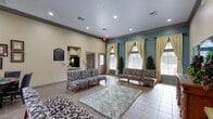 This single-floor clubhouse suite is thoughtfully designed to serve as a welcoming communal gathering space. The living room is the heart of the suite, characterized by elevated ceilings, recessed lighting, and a two-tone color scheme pairing warm cream walls with a rich teal accent wall. Three large windows dressed with elegant cream and gold curtains flood the room with natural light, offering views of the surrounding outdoor areas. Decorative topiary plants flanking the windows add a refined, classic touch. Seating is plentiful, with multiple patterned sofas and armchairs arranged around a soft area rug, creating both intimate conversation nooks and open gathering areas. A carved wooden console along one wall supports a wall-mounted flat-screen television, flanked by abstract canvas artwork. The kitchen, accessible via a pass-through window from the living area, features white cabinetry, generous counter space, and appliances including a microwave and toaster oven. Ornamental iron wall décor adds character to the kitchen's neutral backdrop. The suite is completed by tile flooring throughout, crown molding details, and a dedicated dining area, making it a versatile and inviting space for a variety of events and social gatherings.