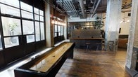 This single-floor space showcases a compelling industrial aesthetic, blending raw architectural character with inviting social amenities. Soaring ceilings reveal exposed pipes and ductwork overhead, while original brick columns and large steel-framed windows allow generous natural light to pour in across the richly worn hardwood floors. A full-length shuffleboard table takes center stage, making it an ideal gathering spot for casual entertainment. Adjacent to the game area, a substantial bar counter finished in dark metal and warm wood tones serves as the focal hub of the space, lined with sturdy industrial-style bar stools. Pendant Edison bulb fixtures hang in clusters, casting a warm, ambient glow that complements the moody, loft-like atmosphere. Lush potted greenery softens the hard edges of the industrial elements, adding life and texture to the overall environment. The steel-framed interior windows and partitions further reinforce the converted-warehouse character of the space. Whether used as an event venue, a social club, or a hospitality destination, this space offers a sophisticated yet laid-back ambiance that balances rugged industrial design with comfortable, curated details throughout.