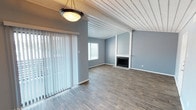 This single-story space offers a thoughtfully designed layout with a variety of architectural details that give it a distinctive character. The living and dining areas are united by a striking white wood-planked ceiling with exposed beams, adding warmth and visual interest overhead. Cool gray walls paired with wood-style flooring create a cohesive, modern aesthetic throughout these shared spaces. A fireplace with a clean white surround serves as a focal point in the living area, while vertical blinds covering a sliding glass door and an additional window allow for flexible natural light control. A pendant light fixture adds a decorative touch to the dining area.

The kitchen is efficiently arranged with white cabinetry featuring sleek bar-pull hardware, light countertops, a blue subway tile backsplash, and stainless steel appliances including a range, microwave, and dishwasher. A double sink and a pantry with bifold doors add practical functionality.

The bedroom features plush carpet, a window with natural light, and direct access to a vanity area with white cabinetry and a large mirror. The bathroom includes a full tub and shower combination with white tile surround. A second carpeted bedroom provides additional flexible living space.