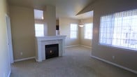 This single-story home presents an open-concept layout connecting the living and dining areas in a seamless, flowing arrangement. The living room is anchored by a white tile-surround fireplace with a clean mantel, serving as a central focal point. Neutral-toned walls and plush wall-to-wall carpeting create a warm, understated aesthetic throughout the space.

Multiple windows are positioned throughout both areas, allowing generous amounts of natural light to fill the interior. Vertical blinds and standard window treatments offer privacy and light control. The dining area is distinguished by a pendant light fixture suspended from above, adding a subtle decorative element to the space.

The vaulted or stepped ceiling detail in the living area adds architectural interest and contributes to a sense of openness. A partial wall divides the living area from an adjoining space, hinting at a kitchen or hallway beyond. White baseboards and trim provide clean contrast against the neutral walls. Overall, the space conveys a comfortable, functional atmosphere suited for everyday living.