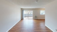 This two-story residence offers a functional and well-appointed layout spread across two levels, connected by an open staircase with a white metal railing. The main level features a spacious living and dining area with warm hardwood-style flooring, textured ceilings, and a wall-mounted HVAC unit. Large sliding glass doors flood the living area with natural light and provide direct access to an outdoor balcony or patio, complemented by a secondary window fitted with blinds. The kitchen is well-equipped, featuring white cabinetry, light quartz countertops, a stainless steel gas range, microwave, dishwasher, and a double-basin sink — offering ample storage and workspace. A bathroom on the main level includes a white vanity with a quartz countertop, a single-basin sink with brushed nickel fixtures, a large mirror with overhead lighting, and clean white finishes throughout. The upper level hosts bedrooms with dark hardwood-style flooring, including rooms with sliding and hinged closet doors providing generous built-in storage. One bedroom features a sliding glass door with outdoor views. The overall aesthetic is clean and neutral, with light gray and beige tones creating a bright, move-in-ready atmosphere throughout the space.