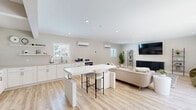 Located at 441 Manor Rd in Newport News, Virginia, this single-floor open-concept space seamlessly blends a fully equipped kitchen with a comfortable and stylishly appointed living area. Light oak-toned luxury vinyl plank flooring runs throughout, lending warmth and continuity to the entire space.

The kitchen is designed for functionality and aesthetics, featuring crisp white shaker cabinetry extending to the ceiling, stainless steel appliances including a French door refrigerator, a deep basin sink beneath a bright window, open floating shelves, and a central white island with bar seating — ideal for casual dining or entertaining.

The living area is open and inviting, highlighted by recessed lighting, a wall-mounted flat-screen TV set within a dark accent media console, and a patterned area rug anchoring the seating arrangement. A marble-look oval coffee table and a pair of curved green velvet accent chairs add a refined, contemporary touch. Natural light fills the room through multiple windows, and a set of glass-paned double doors opens to the outdoors, enhancing the airy ambiance. Split-unit climate control systems are installed throughout, ensuring year-round comfort. The overall aesthetic is clean, modern, and cohesive.